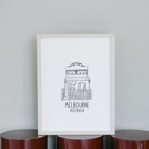 May include: A black and white line drawing of a house with a fence and the text "MELBOURNE AUSTRALIA" underneath. The drawing is framed in a white frame and is sitting on a red cylindrical surface.