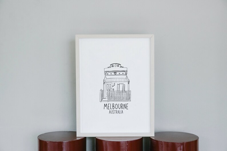 May include: A black and white line drawing of a house with a fence and the text "MELBOURNE AUSTRALIA" underneath. The drawing is framed in a white frame and is sitting on a red cylindrical surface.