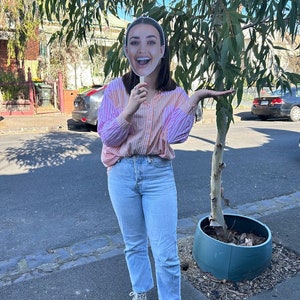 May include: A person holding a cardboard cutout of a woman's face in front of their own face. The cutout has a pink and white striped shirt and a big smile. The person is standing on a sidewalk in front of a tree.