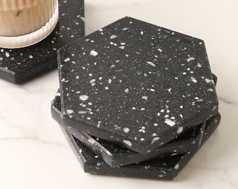 May include: Set of four black and white hexagonal coasters with a speckled pattern. The coasters are stacked on top of each other.