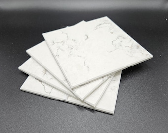 May include: A stack of square white coasters with a marble-like pattern. The coasters have a light gray veining design and are arranged in a fanned-out formation. The coasters are made of a solid material.