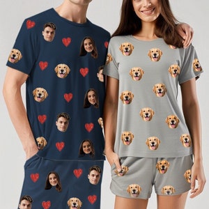 May include: A couple wearing matching pajamas with a repeated pattern of red hearts and faces of a golden retriever dog. The man is wearing a navy blue short-sleeved shirt and matching shorts. The woman is wearing a light gray short-sleeved shirt and matching shorts.