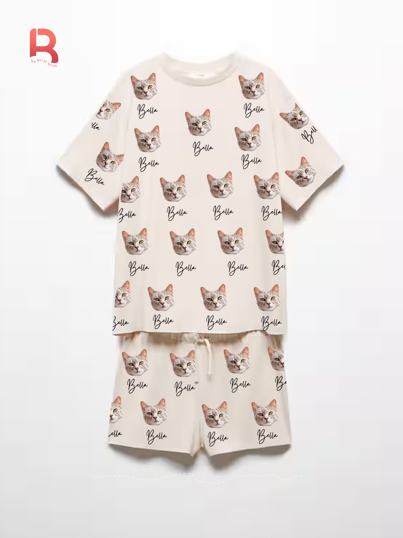 Pu&ograve; includere: Un pigiama color crema con una parte superiore a maniche corte e pantaloncini. Il set presenta un motivo ripetuto di un viso di gatto con il nome "Bella" scritto sotto ogni immagine.
