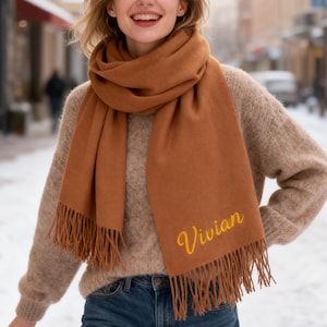 May include: A rust-colored scarf with a fringed edge and the name "Vivian" embroidered in yellow script. The scarf is wrapped around the neck and shoulders. The person is wearing a tan sweater and blue jeans.