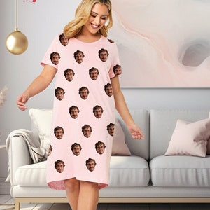 May include: A pink short-sleeved nightgown with a repeating pattern of a man's face. The woman is wearing the nightgown and standing in front of a light grey sofa.