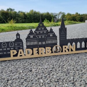Könnte beinhalten: Schwarze Silhouette der Paderborner Skyline mit dem Stadtnamen in goldenen Lettern auf einem Holzsockel.