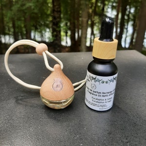 Peut inclure: Un diffuseur d'air pour voiture en bois avec une ficelle pour l'accrocher au rétroviseur. Le diffuseur est rempli d'un liquide clair. À côté se trouve une petite bouteille de recharge de diffuseur d'huile parfumée à l'eucalyptus et à la sauge.