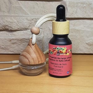 Peut inclure: Un diffuseur d'air pour voiture en bois avec un récipient en verre transparent et une ficelle pour l'accrocher. Le diffuseur d'air est rempli d'un liquide rose et rouge. La bouteille est étiquetée "OR parfum Recharge diffuser, grance Oil Refill diffuser, Explosions de fruits, Fruit burst, paraben free fragrance, Anettatie.com".