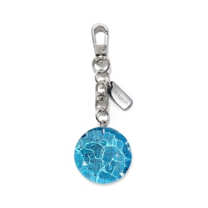 May include: A silver keyring with a round blue and white charm featuring a pool water design. The charm has a silver ring attached to it. The keyring also has a silver tag with the word "Alltaglich" on it.