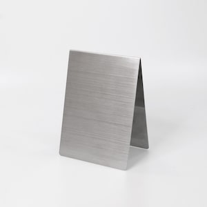 May include: A V-shaped table sign holder made of brushed stainless steel with a metallic silver finish. The object features a rectangular front panel with a visible horizontal grain texture and a folded design that allows it to stand upright.