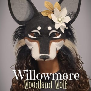 Puede incluir: Una máscara de lobo negra, blanca y marrón con orejas grandes y puntiagudas, cubiertas de malla para los ojos y un adorno floral con hojas blancas y doradas. El texto Willowmere Woodland Wolf y @CheetahPawsCreations está impreso en la imagen.