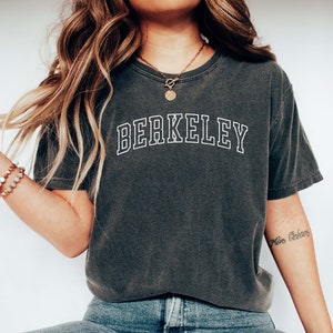 May include: A dark gray t-shirt with the word "BERKELEY" printed in white outlined letters. The person is wearing blue jeans and white sneakers.