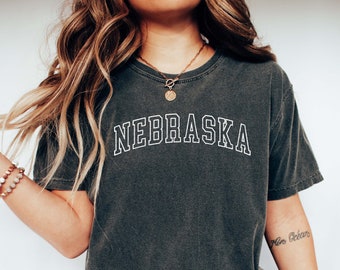 May include: A woman wearing a dark gray t-shirt with the word "NEBRASKA" printed in white on the front. She is also wearing blue jeans and white sneakers.