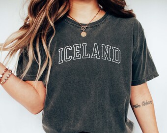 May include: A dark gray t-shirt with the word "ICELAND" printed in white outlined letters. The person wearing the shirt is also wearing blue jeans and white sneakers.