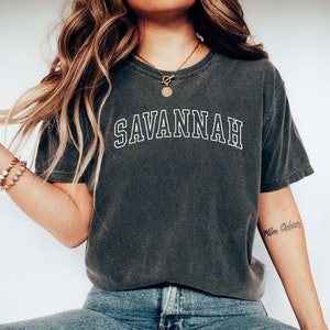 May include: Dark gray t-shirt with the word "SAVANNAH" in white, collegiate-style lettering. The shirt is paired with blue jeans and white slip-on shoes. A gold necklace and bracelets are also visible.