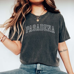 May include: A woman wearing a dark gray t-shirt with the word "PASADENA" printed in white on the front. She is also wearing blue jeans and white sneakers.