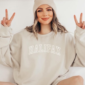 May include: A beige sweatshirt with the word "HALIFAX" printed in white on the front. The sweatshirt has a crew neck and long sleeves.