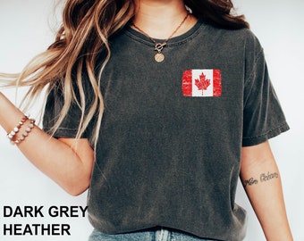 May include: A dark grey heather t-shirt with a distressed Canadian flag graphic on the left chest. The model is wearing blue jeans and white sneakers.