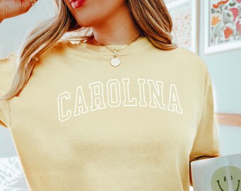May include: A yellow t-shirt with the word "CAROLINA" printed in white on the front. The shirt is being worn by a person with blonde hair and a gold necklace.