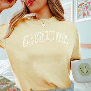 Pode incluir: Um moletom amarelo claro com a palavra "HAMILTON" em letras brancas. A pessoa est&aacute; segurando uma caneca com um rosto sorridente e um livro intitulado "How to Plant House Plants".
