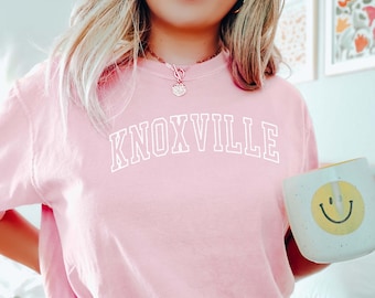 May include: A pink t-shirt with the word "KNOXVILLE" printed in white on the front. The person is wearing blue shorts and holding a white mug with a yellow smiley face.