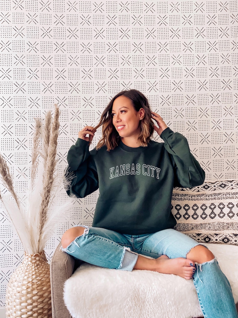 K&ouml;nnte beinhalten: Eine Frau tr&auml;gt ein dunkelgr&uuml;nes Sweatshirt mit dem Schriftzug "KANSAS CITY" in wei&szlig;en Buchstaben. Sie sitzt auf einem wei&szlig;en Sofa mit einer wei&szlig;en Wolldecke und einem gemusterten Kissen. Sie tr&auml;gt eine blaue Jeans mit Rissen an den Knien.