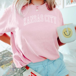 Op de afbeelding: Een roze T-shirt met de tekst "KANSAS CITY" in witte letters. Het shirt wordt gedragen door een persoon en wordt gecombineerd met een lichtblauwe short.
