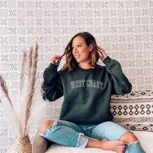 May include: A woman wearing a dark green sweatshirt with the text "WEST COAST" in white lettering. She is sitting on a white couch with a patterned pillow behind her. The background is a white wall with a black and white geometric pattern.