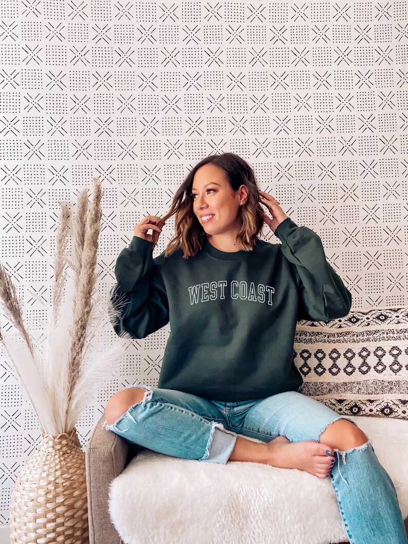 May include: A woman wearing a dark green sweatshirt with the text "WEST COAST" in white lettering. She is sitting on a white couch with a patterned pillow behind her. The background is a white wall with a black and white geometric pattern.