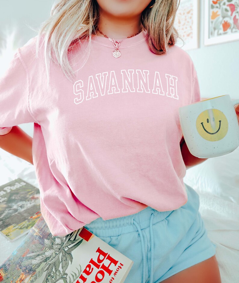 May include: A light pink sweatshirt with the word "SAVANNAH" in white block letters. A white mug with a yellow smiley face is held in the hand. The person is wearing light blue shorts.