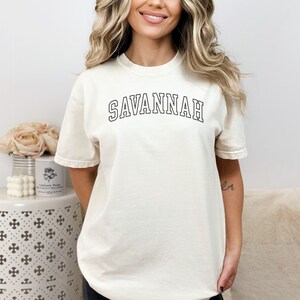 May include: Cream-colored t-shirt with the word "SAVANNAH" in black, arched lettering. The shirt has short sleeves and a relaxed fit. The text is outlined in black. The shirt is made of a soft, comfortable material.