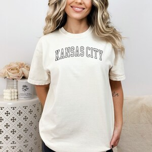 Op de afbeelding: Een wit T-shirt met de tekst "KANSAS CITY" in zwarte omrande letters.