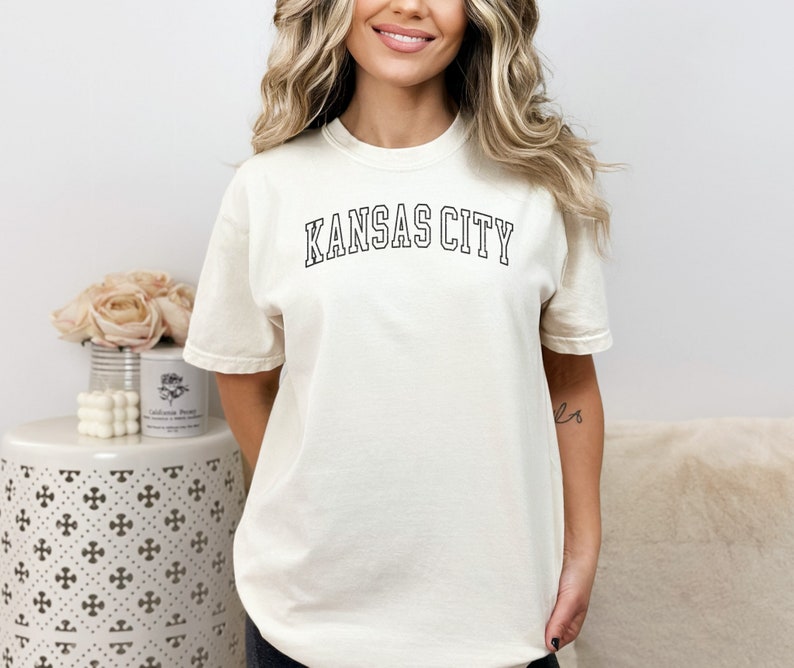 Op de afbeelding: Een wit T-shirt met de tekst "KANSAS CITY" in zwarte omrande letters.