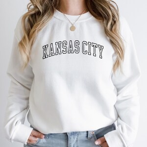 K&ouml;nnte beinhalten: Wei&szlig;er Pullover mit schwarzem Text, der "KANSAS CITY" in einer College-Schriftart lautet.