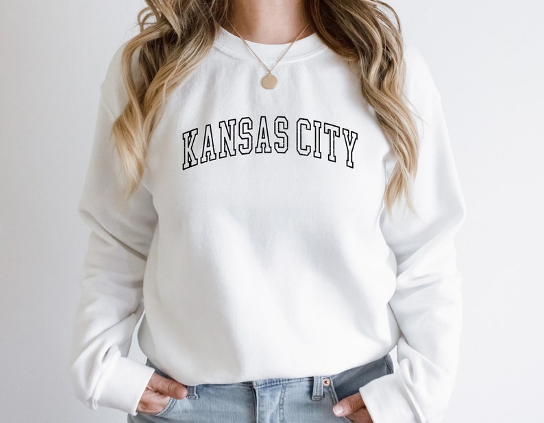 K&ouml;nnte beinhalten: Wei&szlig;er Pullover mit schwarzem Text, der "KANSAS CITY" in einer College-Schriftart lautet.