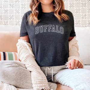 May include: A woman wearing a dark gray t-shirt with the word "BUFFALO" in white lettering. She is sitting on a white couch with a white crocheted cardigan draped over her shoulders.