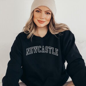 May include: A black sweatshirt with the word "NEWCASTLE" printed in white on the front. The sweatshirt has a crew neck and long sleeves.