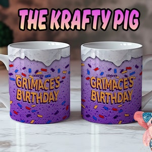 May include: Two white ceramic mugs with purple and yellow designs. The mugs have the text "Celebrate Grimace's Birthday" on them. The mugs are on a white marble surface. A pink pig with a blue bow is holding a donut in the bottom right corner of the image.