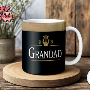 以下が含まれることがあります： 白い取っ手を持つ黒いセラミックマグカップ。マグカップには、竪琴と「20 24 GRANDAD」という文字が書かれた金色のデザインが施されています。
