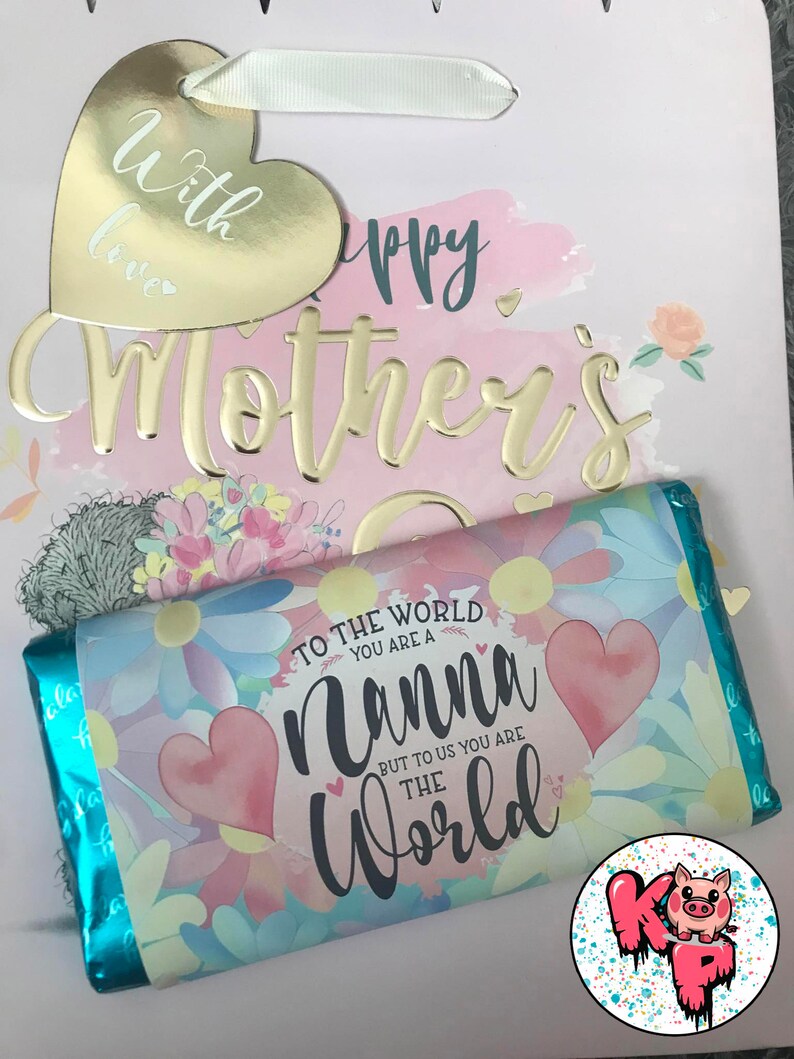 Pode incluir: Uma sacola de presente rosa e azul com uma etiqueta de cora&ccedil;&atilde;o dourada que diz "With love" e uma inscri&ccedil;&atilde;o dourada que diz "Happy Mother's Day". A sacola cont&eacute;m uma barra de chocolate com um design floral e o texto "To the world you are a Nanna but to us you are the World".