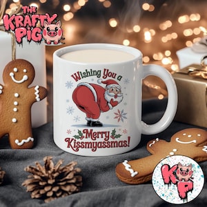 Può includere: Tazza in ceramica bianca con manico, con una grafica di Babbo Natale che si china, con la scritta "Wishing You a Merry Kissmyassmas!". La tazza è circondata da decorazioni natalizie, tra cui un omino di pan di zenzero e una pigna.