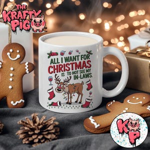 Op de afbeelding: Witte keramische mok met de tekst "ALL I WANT FOR CHRISTMAS IS TO NOT SEE MY IN-LAWS." De mok heeft een cartoon rendier, kerstsokken en wijnglazen. Een peperkoekmannetje en een dennenappel staan ook op de foto.