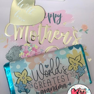 Pode incluir: Uma sacola de presente com uma etiqueta de cora&ccedil;&atilde;o dourada que diz "with love" e uma inscri&ccedil;&atilde;o em folha de ouro que diz "Happy Mother's Day". A sacola cont&eacute;m uma barra de chocolate com um design floral e o texto "World's Greatest Grandma".