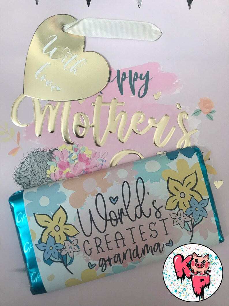 Pode incluir: Uma sacola de presente com uma etiqueta de cora&ccedil;&atilde;o dourada que diz "with love" e uma inscri&ccedil;&atilde;o em folha de ouro que diz "Happy Mother's Day". A sacola cont&eacute;m uma barra de chocolate com um design floral e o texto "World's Greatest Grandma".