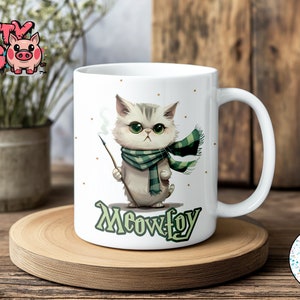 May include: White ceramic mug with a cartoon illustration of a cat wearing a green and white scarf and holding a wand. The cat has green eyes and a grumpy expression. The mug has the text "Meowfoy" printed on the bottom.