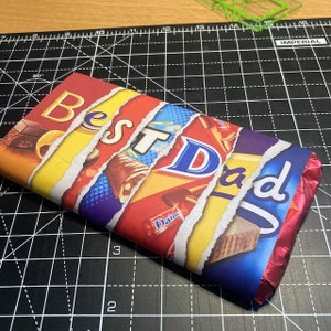 Pode incluir: Uma barra de chocolate embrulhada em papel alum&iacute;nio colorido com as palavras "Best Dad" impressas nela. O papel alum&iacute;nio est&aacute; rasgado em alguns lugares para revelar as barras de chocolate que est&atilde;o por baixo.