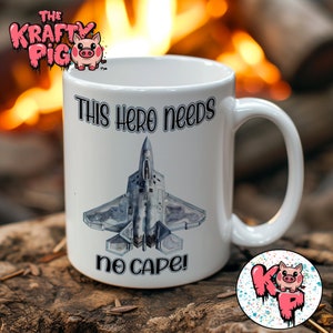 May include: White ceramic mug with a black and gray fighter jet graphic and the text "This hero needs no cape!" printed on it. The mug is sitting on a wooden surface.