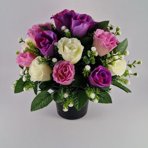 May include: A bouquet of artificial flowers in a black pot. The flowers are pink, white, and purple roses with small white flowers interspersed throughout.