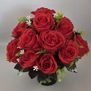 May include: A bouquet of vivid red roses, arranged with green leaves and small white flowers. The roses are in full bloom, creating a lush and romantic display. The arrangement is set in a dark vase, offering a striking contrast.