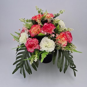 May include: A pink and white carnation flower arrangement in a black pot. The arrangement features green leaves and small white flowers.